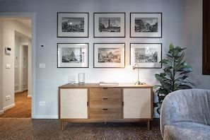A room with a wooden cabinet, six framed black-and-white cityscape photographs on the wall, a lamp, and a large green plant. - Ca' Vida Piano Nobile (Venezia)