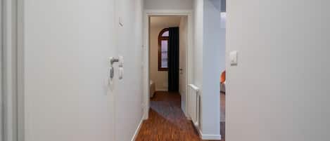 Un pasillo con suelo de madera, paredes blancas y una puerta que da a otra habitación.