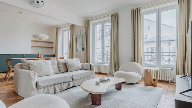 A modern living room with a sofa, armchair, and coffee table. There is a window with curtains and a view of buildings outside. - Blueground | Bastille, near shops & park (Paris)