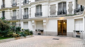 Un patio con suelo empedrado, la entrada de un edificio con puertas de cristal y una pequeña zona ajardinada con plantas.