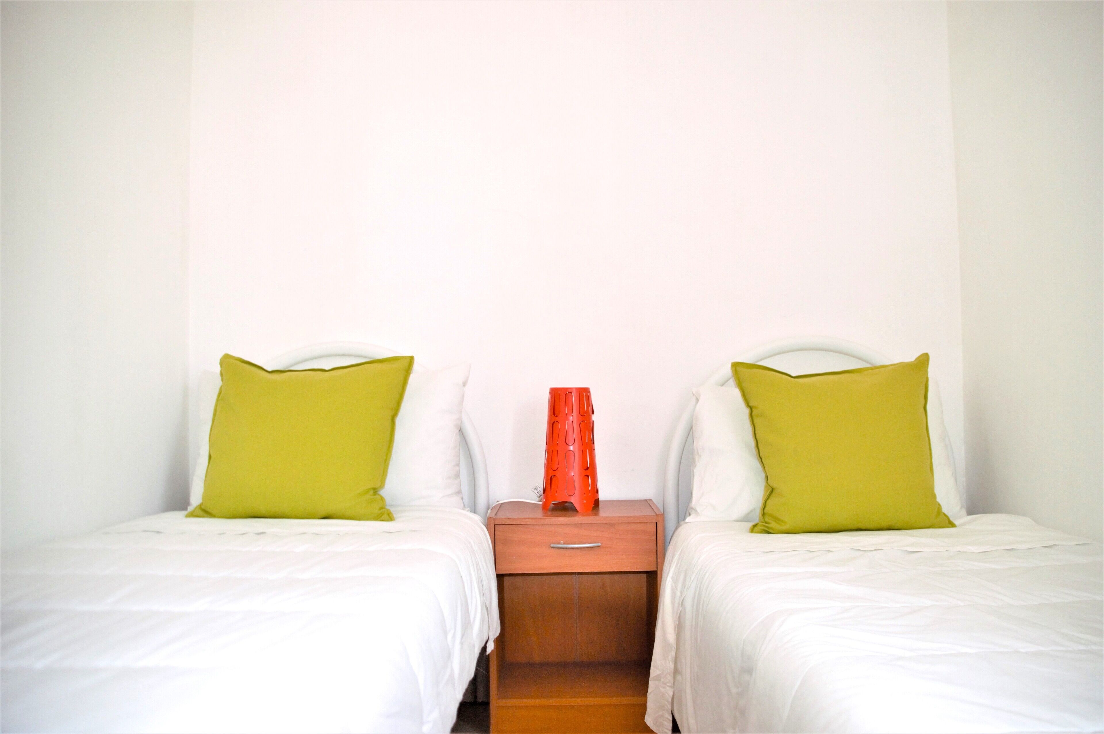 Una habitación con dos camas individuales, cada una con ropa de cama blanca y una almohada verde. Hay una mesita de noche de madera con un jarrón rojo entre las camas.