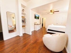 A bedroom with a bed, a chair, a dresser, a mirror, a ceiling fan, and a TV.