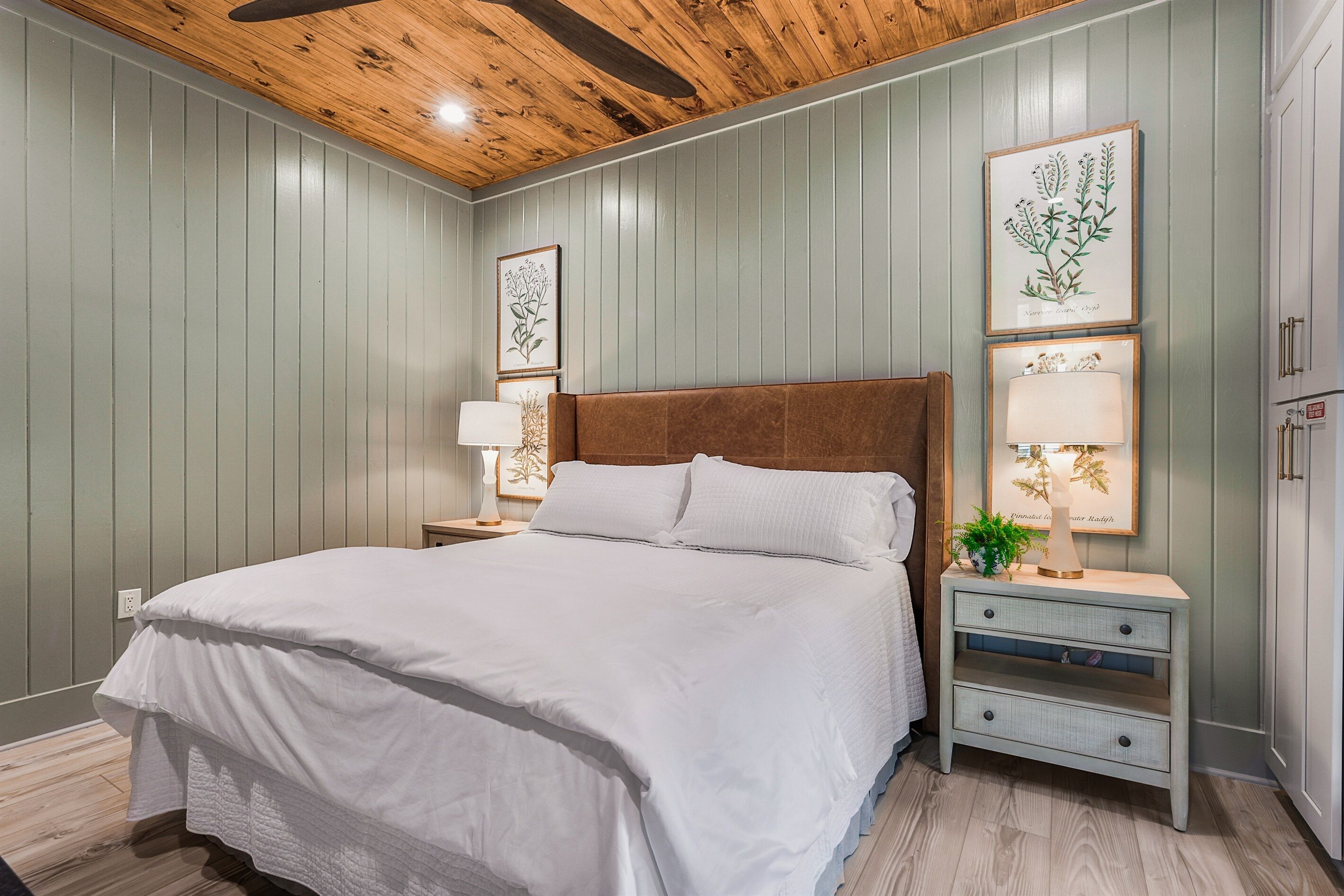 Une chambre avec un plafond en bois, un lit recouvert de linge de lit blanc, une table de chevet avec une lampe, et des reproductions encadrées de motifs botaniques accrochées au mur.