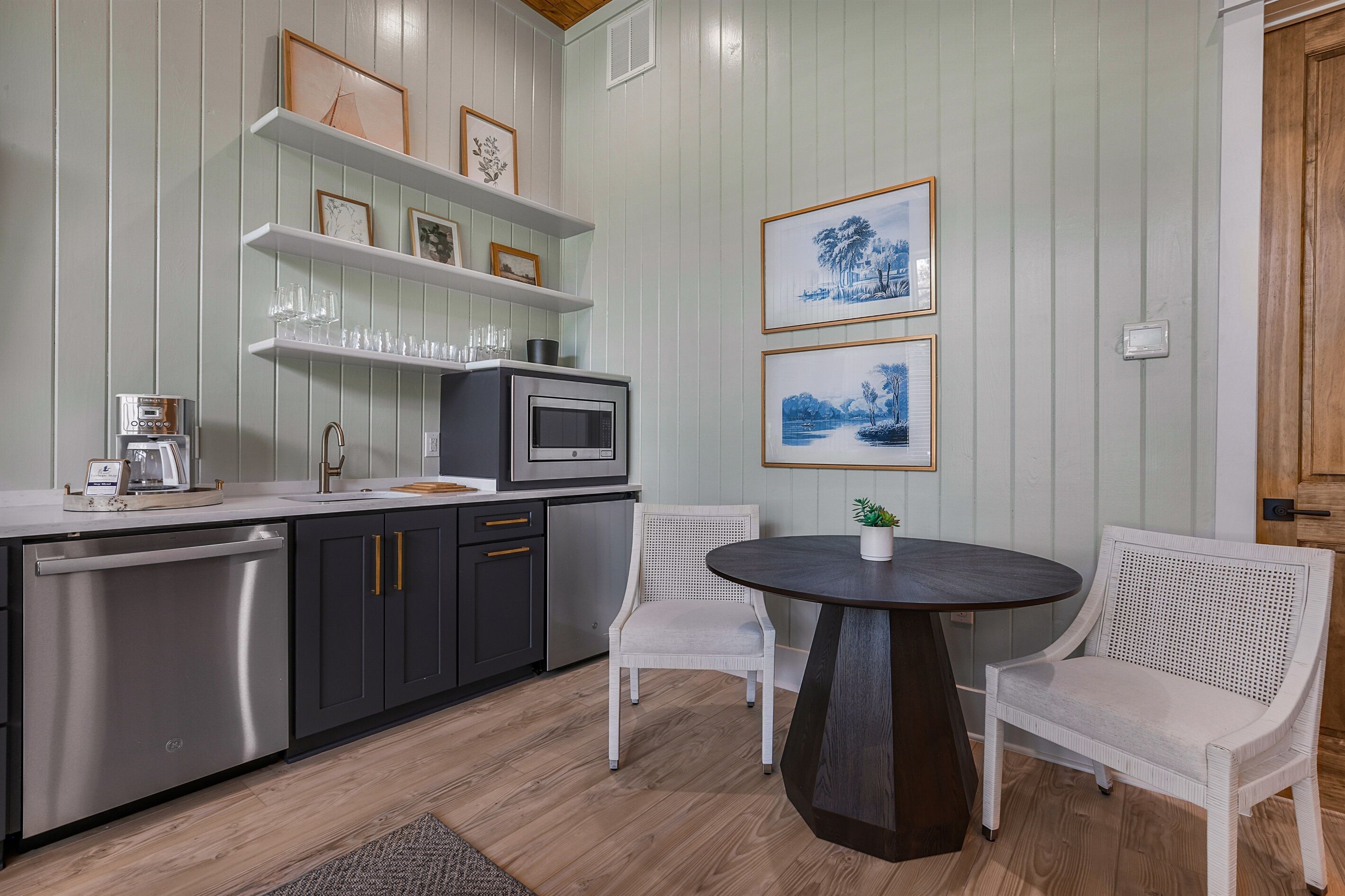 A modern kitchen with stainless steel appliances, white wicker chairs, and framed artwork on the wall.