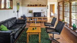 Un soggiorno con un divano in pelle nera, un tavolino in legno e una poltrona in pelle nera. Si nota una libreria con libri e oggetti decorativi, una televisione e una finestra con vista sul verde.