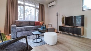 Ein modernes Wohnzimmer mit einem grauen Sofa, einem kleinen runden Tisch, einem weißen Pouf, einem Fernseher auf einem Holzschrank und einem großen Fenster mit Vorhängen.