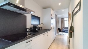 Eine moderne Küche mit schwarzem Arbeitsplatz, weißen Schränken und einem eingebauten Backofen. Ein Fenster mit Jalousien sowie ein Spülbecken mit Armatur sind vorhanden. Im Hintergrund sind ein Stuhl und eine Tasche zu sehen.