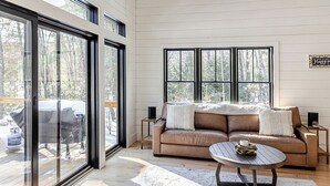 A cozy living room with a beige sofa, a round coffee table, and large windows offering a view of the outdoors. - Lakefront, brand new, craftsman style 4-bedroom 2 bath house home on Long Lake. (Bridgton)