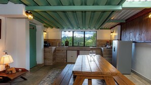 A rustic kitchen with a wooden table, a refrigerator, and a window offering a view of greenery.
