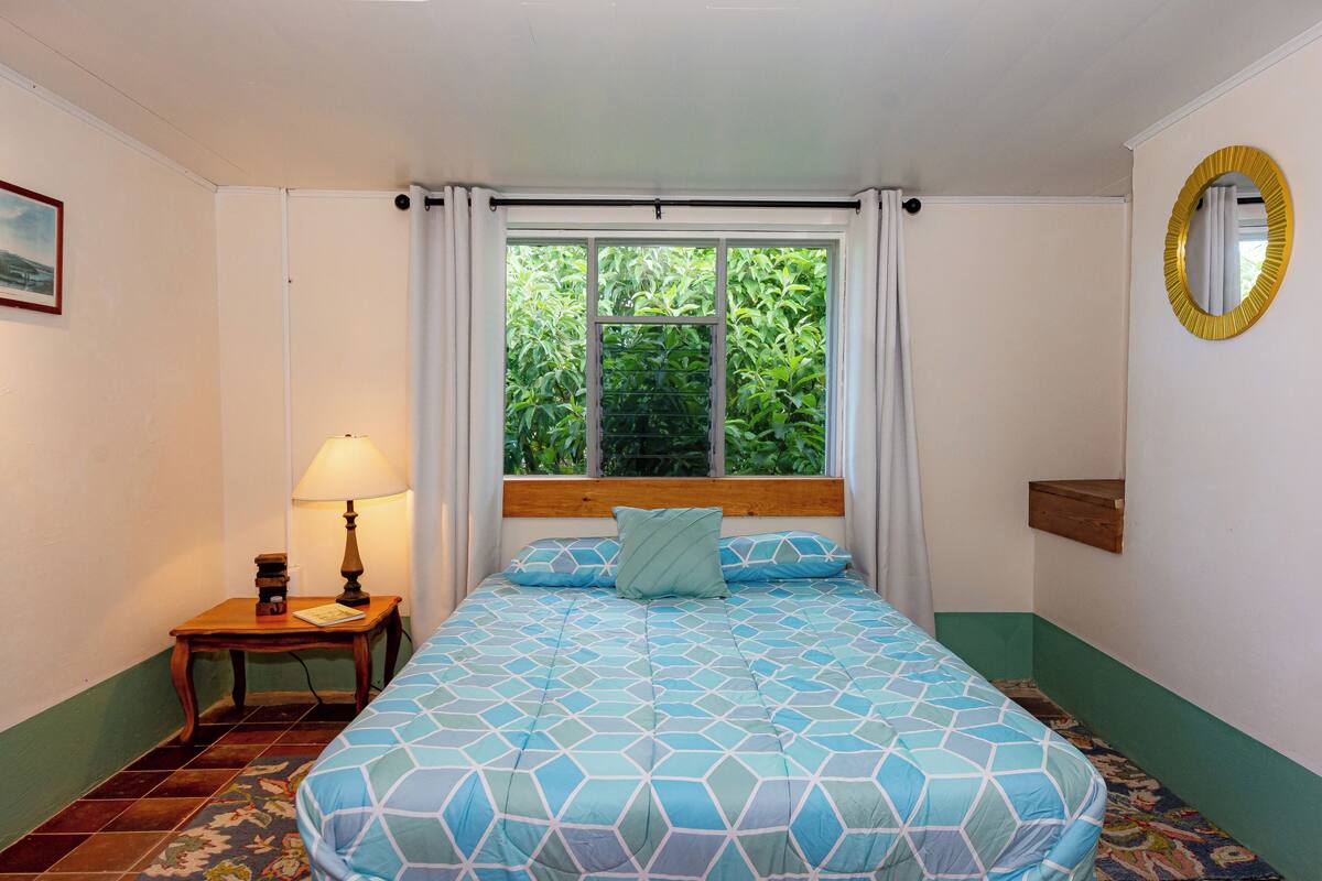 Un dormitorio con cama, mesita de noche, lámpara, ventana con cortinas y espejo en la pared.