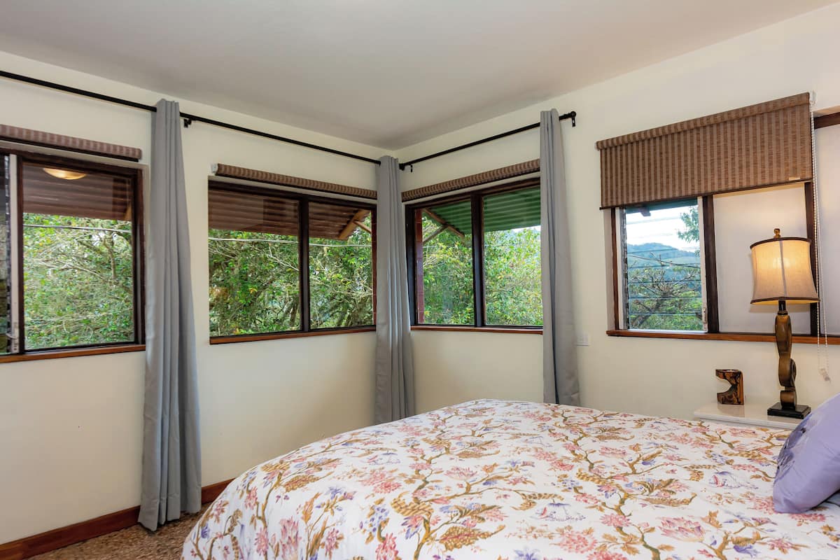 Un dormitorio con cubrecama floral, ventanas amplias con cortinas y una lámpara en la mesita de noche.