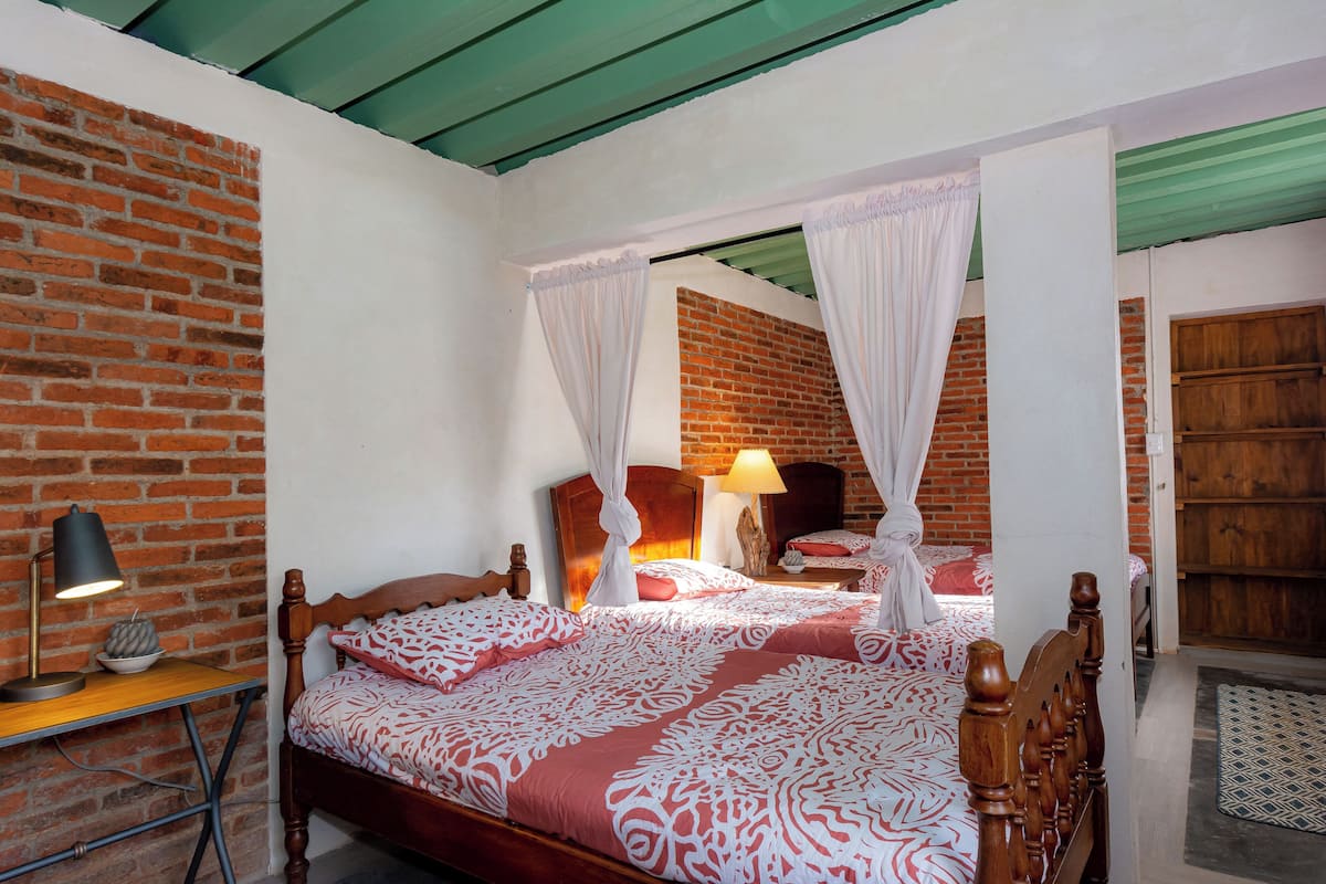 Una habitación con pared de ladrillo, una cama de madera con ropa de cama blanca y roja, una mesita de noche con lámpara y un armario de madera.
