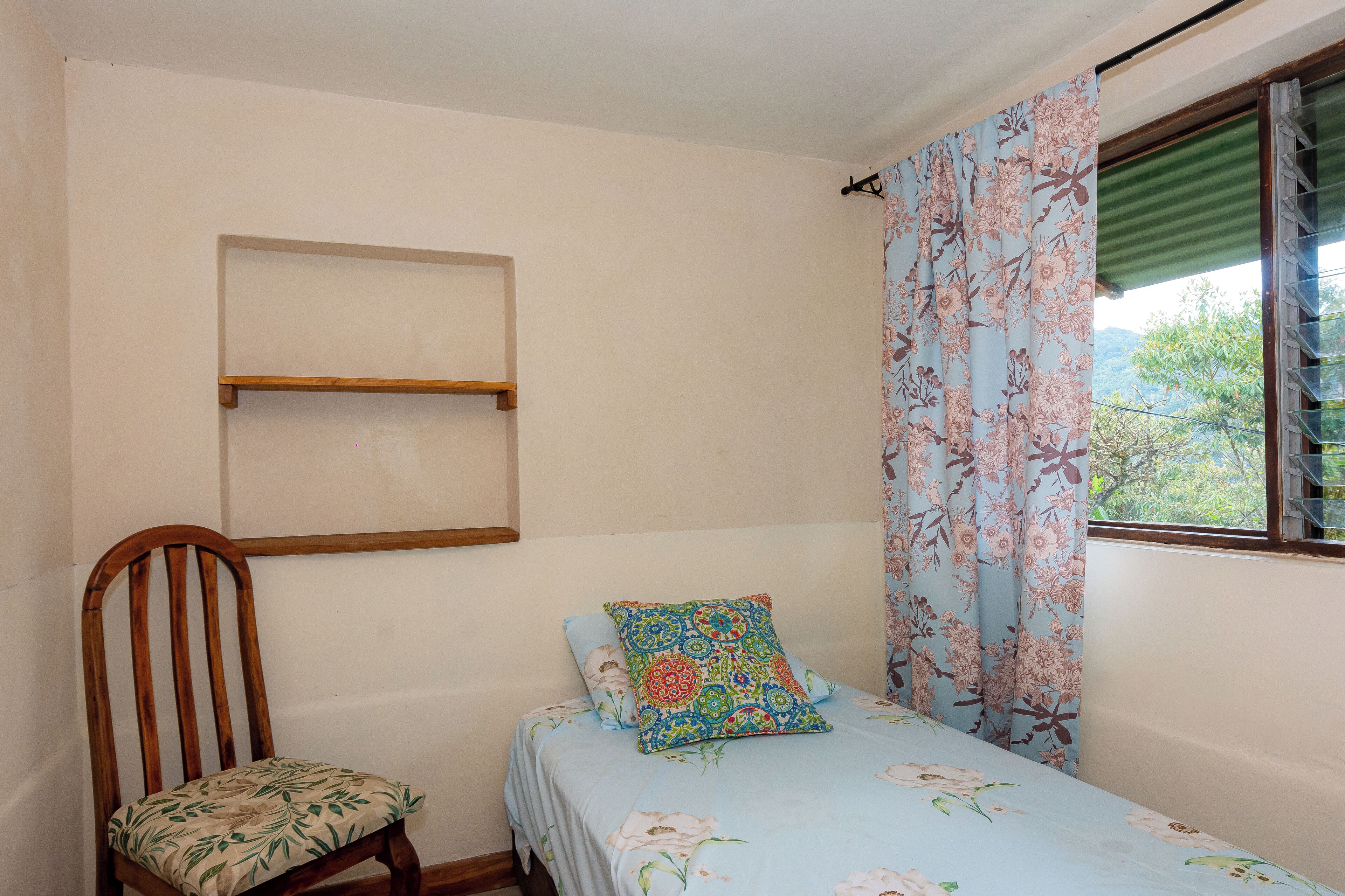 A small bedroom with a single bed, a wooden chair, a shelf, and a window with floral curtains.