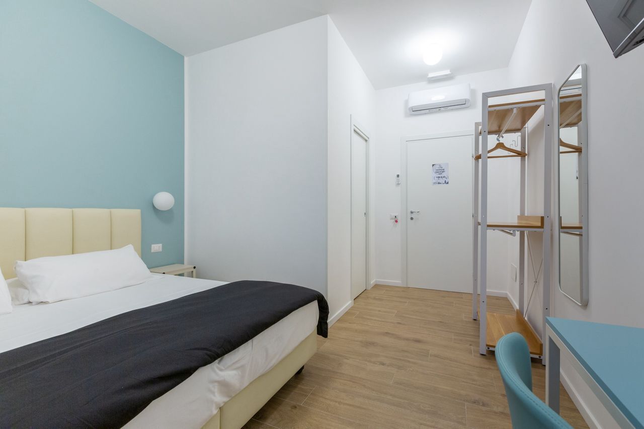 A bedroom with a bed, a desk, and a chair. There is a wardrobe with a mirror and a hanging rack.