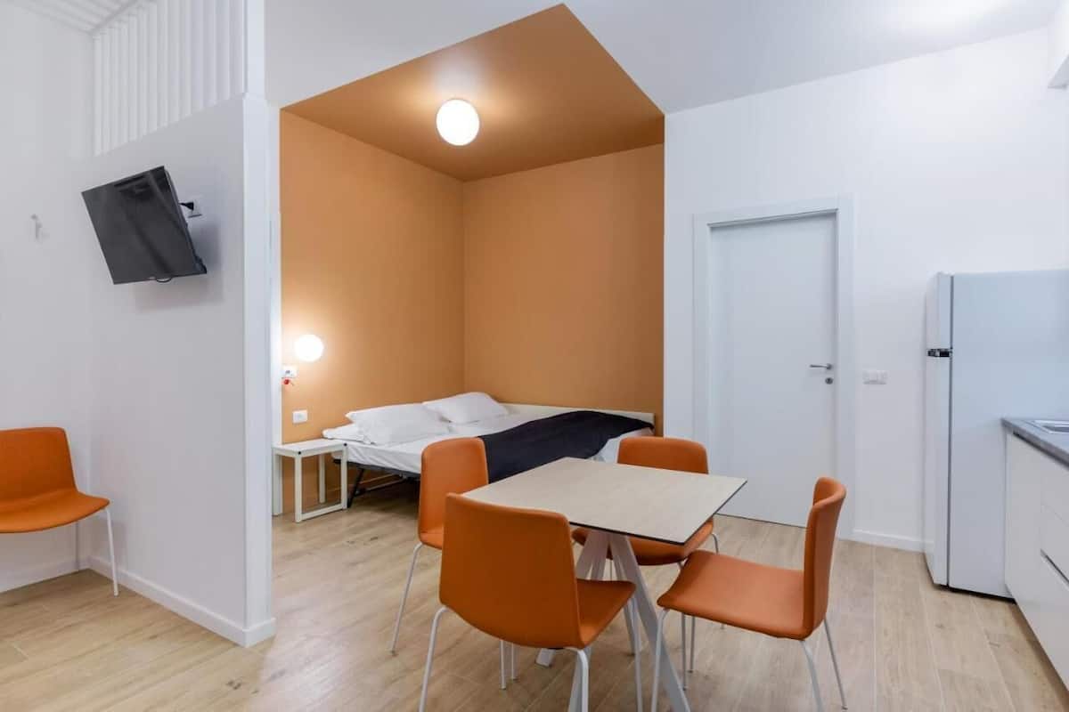 A compact room with a bed, a small table, and orange chairs. There is a TV mounted on the wall and a door leading to another room.