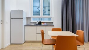 Una cuina compacta amb un frigorífic blanc, una finestra amb reixes decoratives, un rentaplat i una petita encimera. Hi ha una taula amb quatre cadires taronja i una cortina grisosa a la dreta.