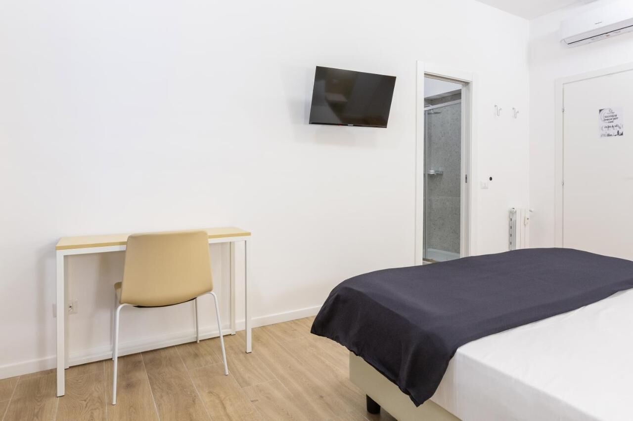 A bedroom with a bed, a desk with a chair, a television mounted on the wall, and a door leading to a bathroom.