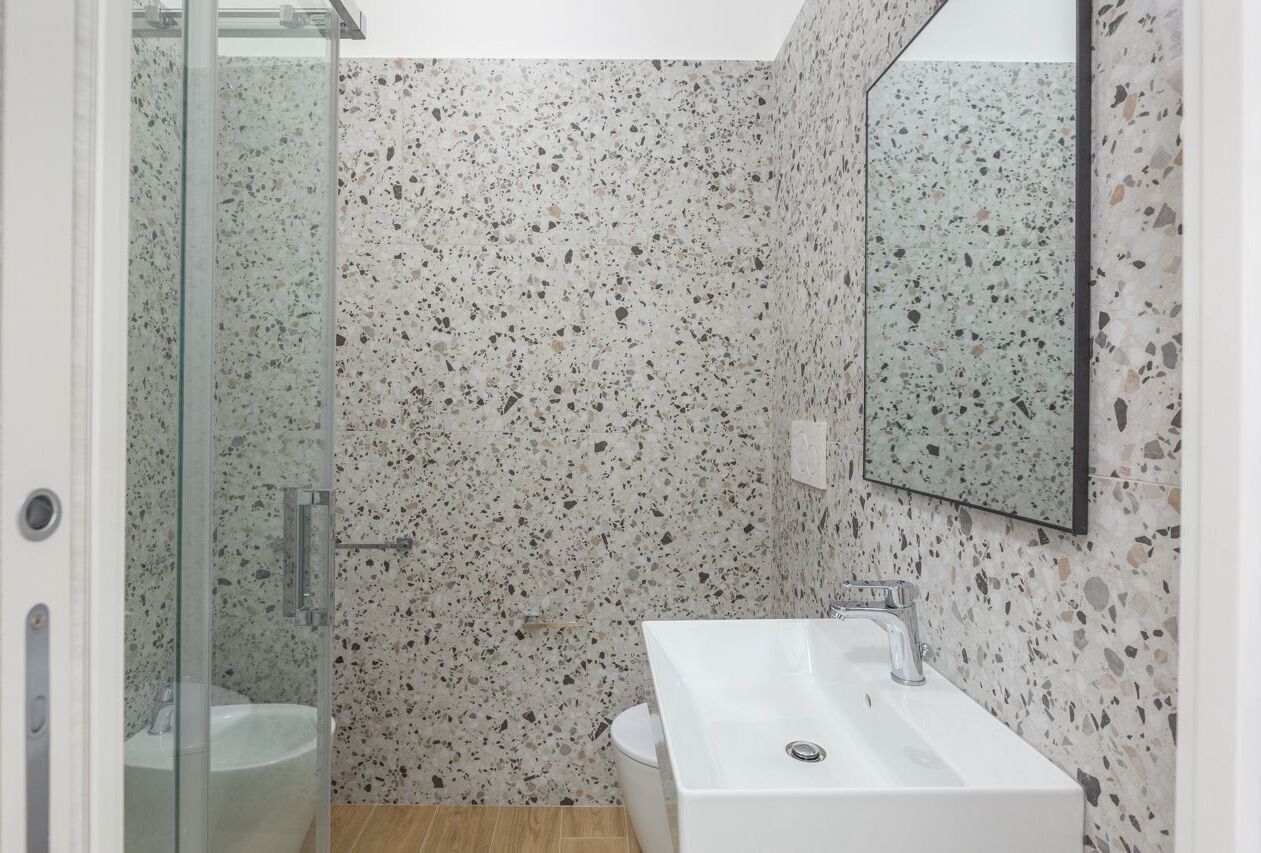 Un baño con un lavamanos blanco, cabina de ducha con mampara de vidrio y pared de azulejos con vetas.