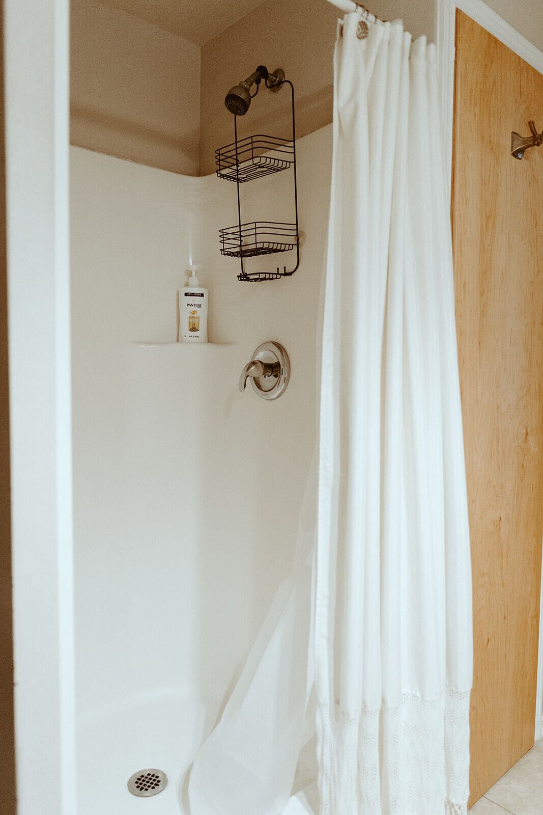 A bathroom with a shower, a towel rack, a soap dispenser, and a wooden door.