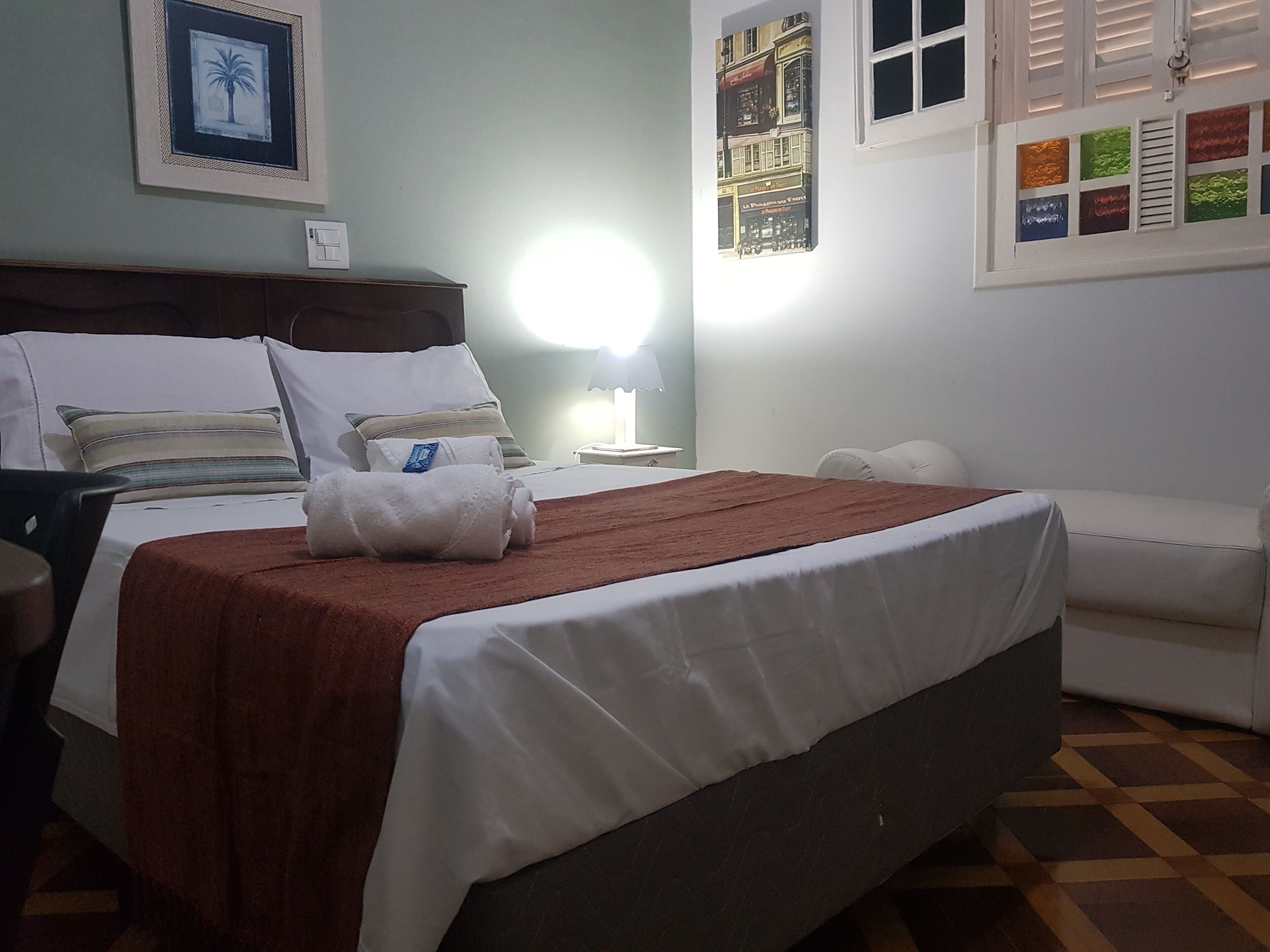 Una cama bien hecha con una manta marrón y almohadas blancas, una mesita de noche con una lámpara, y una pared con cuadros enmarcados.