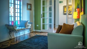 A hotel lobby with a green wall, a white sofa, a bench with striped cushions, a blue rug, and a door marked "SAÍDA".