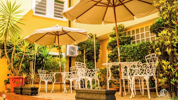 An outdoor patio with white metal chairs and tables, surrounded by potted plants and umbrellas.