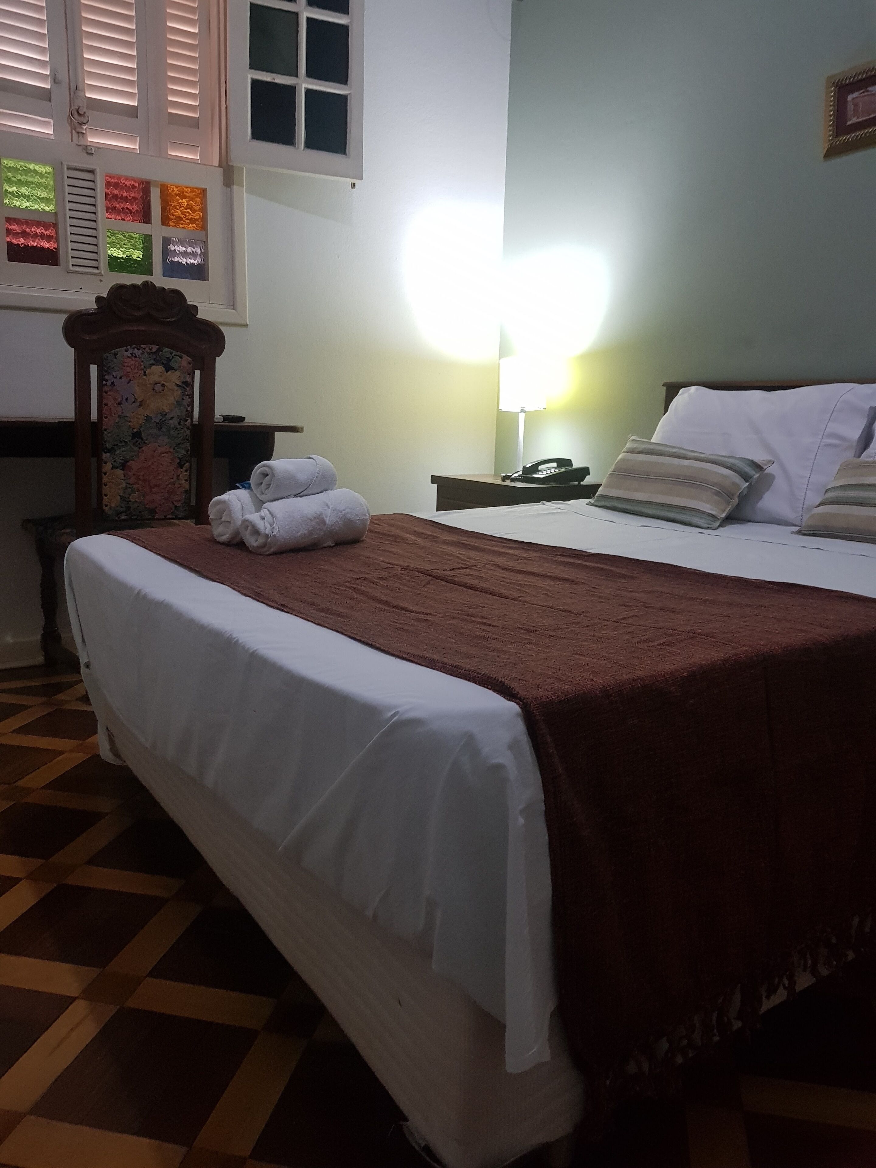 Habitación de hotel con cama, mesita de noche, lámpara y ventana con persianas.