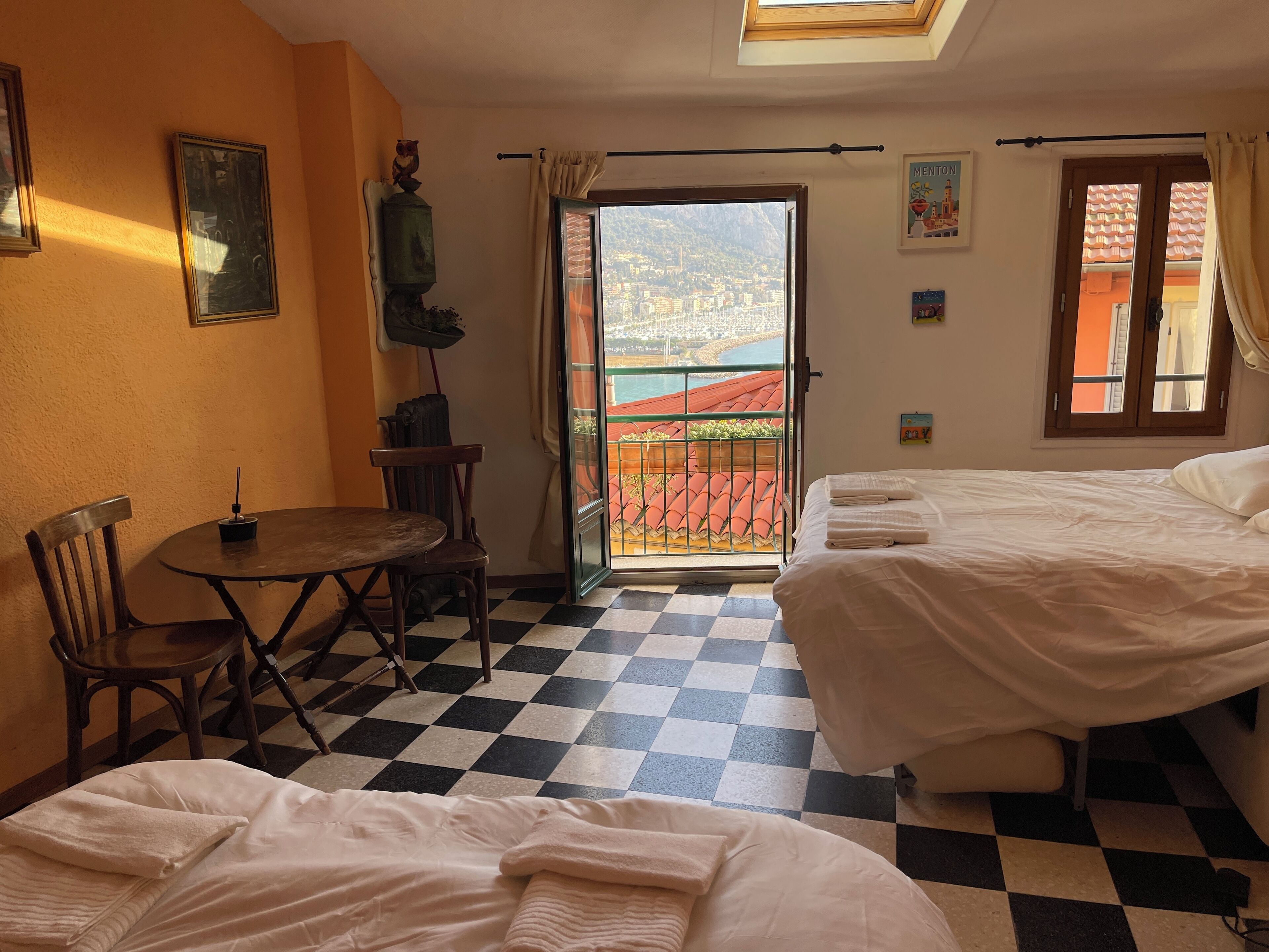 Una habitación de hotel con piso a cuadros, dos camas, una mesa con sillas y un balcón con vista a la ciudad.