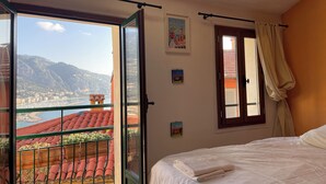 A bedroom with a bed, a window with curtains, a balcony with red tiles, and a view of a coastal town. - House Old town 50m beach'shop (Menton)