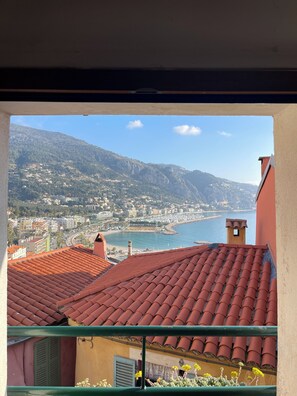 Un balcon doté d’un toit recouvert de tuiles rouges, surplombant une ville côtière et offrant une vue dégagée sur la mer et les montagnes.