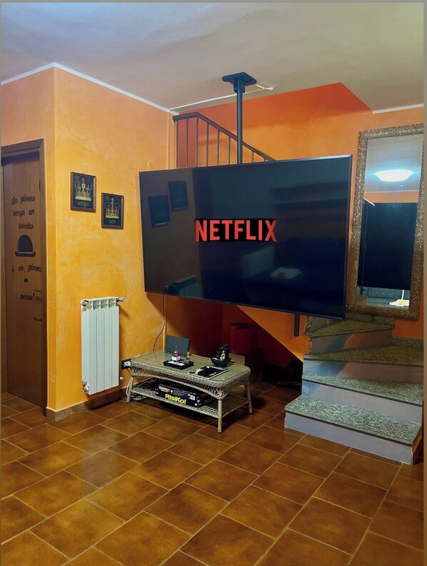 Un salon doté d’un grand téléviseur à écran plat fixé au mur, d’un escalier avec un miroir et d’un radiateur posé sur le sol.