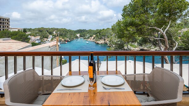 Un balcón con una mesa lista para dos personas, con vista a la costa y al mar, rodeado de vegetación.
