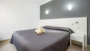 En pænt redt seng med et sammenrullet håndklæde, et natbord og en vægmonteret lampe.