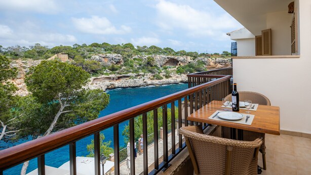 En balkon med et bord dækket til to, med udsigt over en klippefyldt kystlinje og krystalklart, blåt hav.