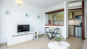 En moderne stue med et fladskærms-tv, et spiseområde med stole, et lille bord med en plante, og et køkken med skabe og hårde hvidevarer.
