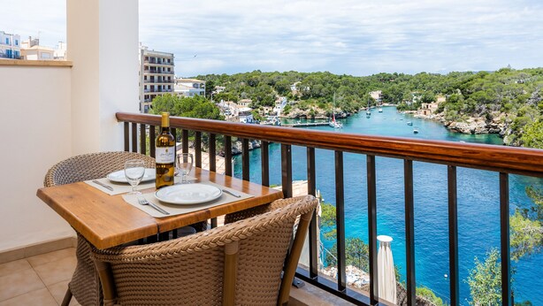 Un balcon avec une table en bois, dressée pour le dîner, donnant sur une ville côtière aux eaux bleues et limpides, entourée de verdure.