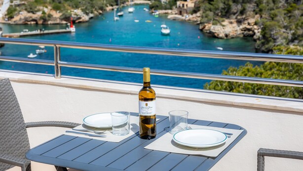 Una mesa con una botella de vino, platos y copas en un balcón con vistas a la costa, con barcos y vegetación.