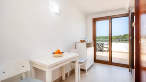 Une petite pièce lumineuse, avec une table blanche, une chaise et un saladier rempli de fruits. Une porte-fenêtre coulissante donne sur un espace extérieur aménagé avec une table et des chaises.