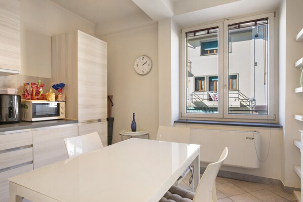 A modern kitchen with a microwave, a dining table, chairs, a clock, and a window with a view of buildings.