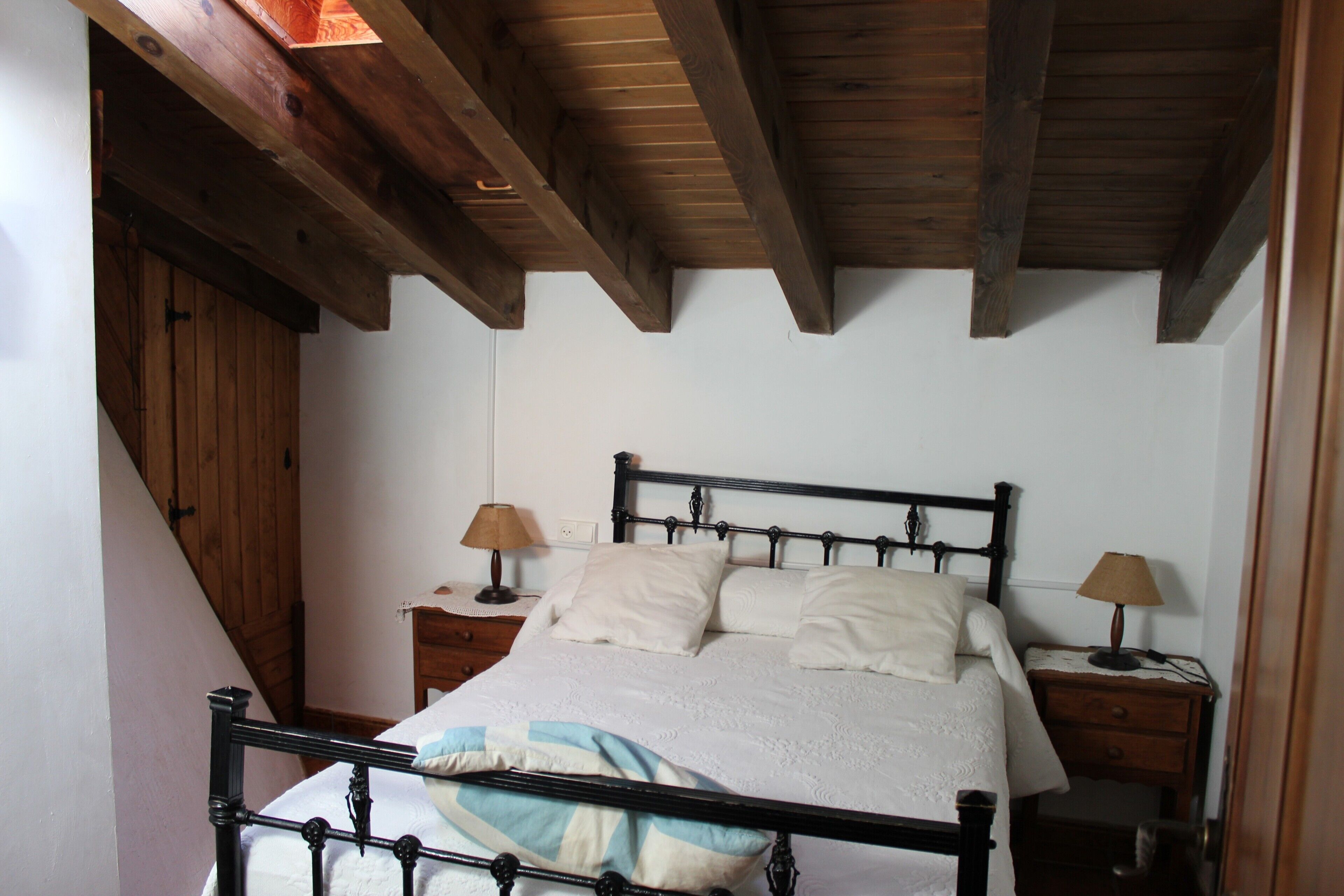 A bedroom with a wooden ceiling, a bed with white bedding, two bedside tables with lamps, and a wooden wardrobe.