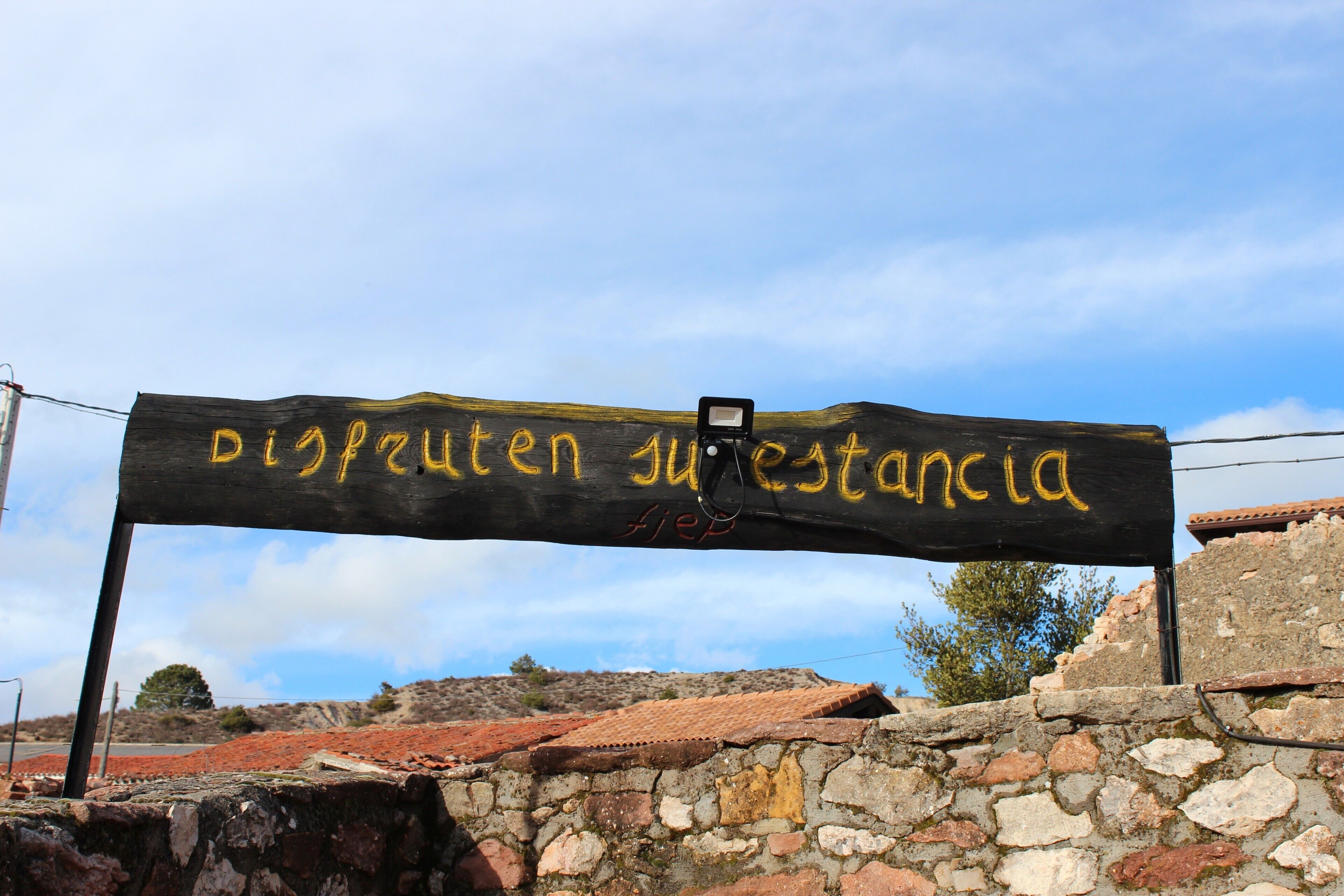 Una pared de piedra rústica con un letrero de madera que muestra el texto 'Disfruten su estadancia' en amarillo.