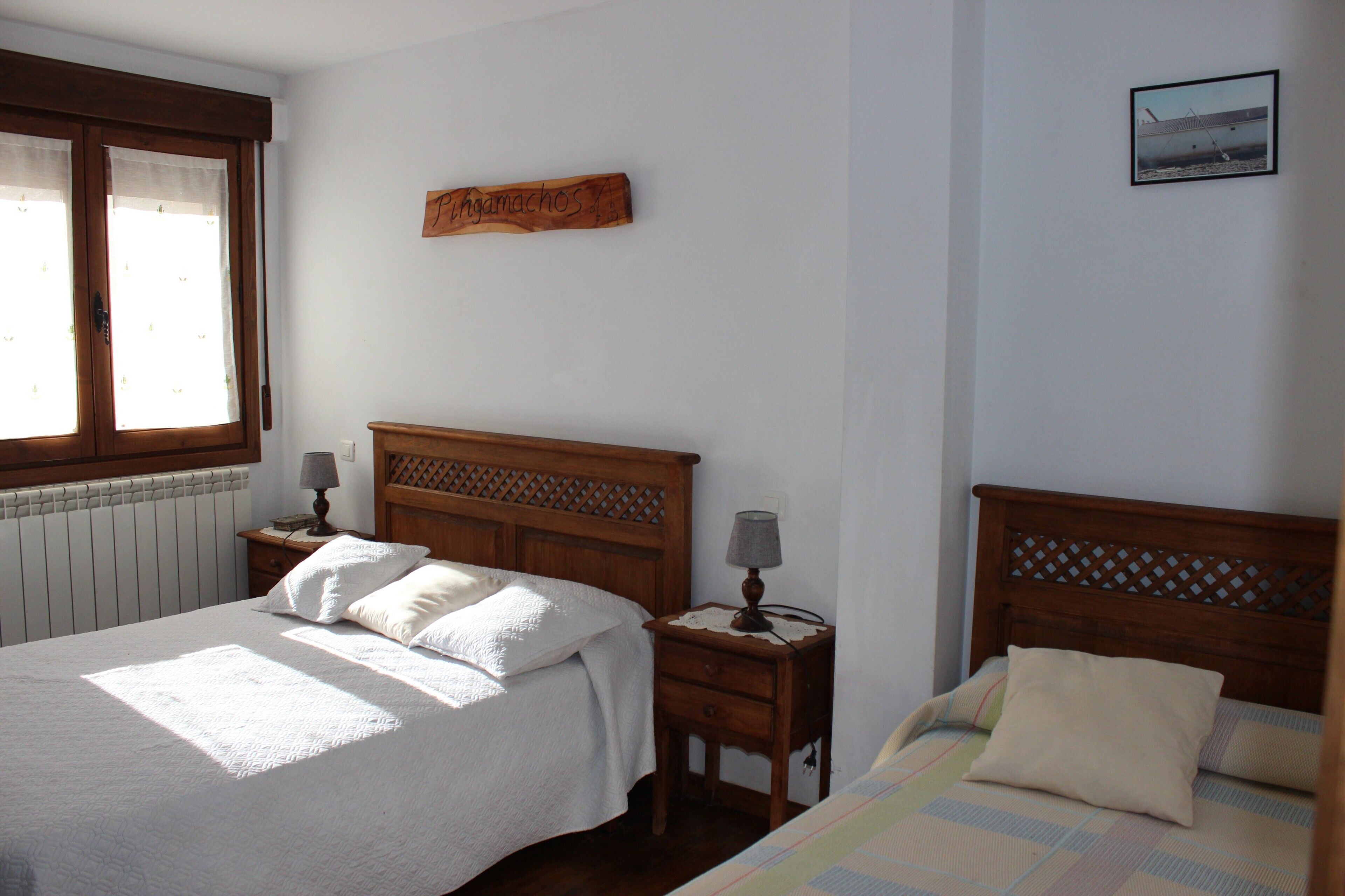 Een slaapkamer met twee bedden, houten hoofdeinden, een nachtkastje met een lamp, een aan de muur hangend kunstwerk en een raam met gordijnen.