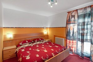 Un dormitorio con cabecera de madera, colcha roja con motivos de ciervos, una mesita de noche y una ventana con cortina que muestra un paisaje de bosque y montaña.