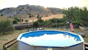 A swimming pool with a slide, surrounded by a wooden fence, and a hill in the background.