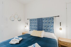 A neatly made bed with a blue bedspread, a white pillow, and a rolled towel on the side. There are two bedside tables with lamps, a wall-mounted mirror, and a patterned tapestry above the bed.