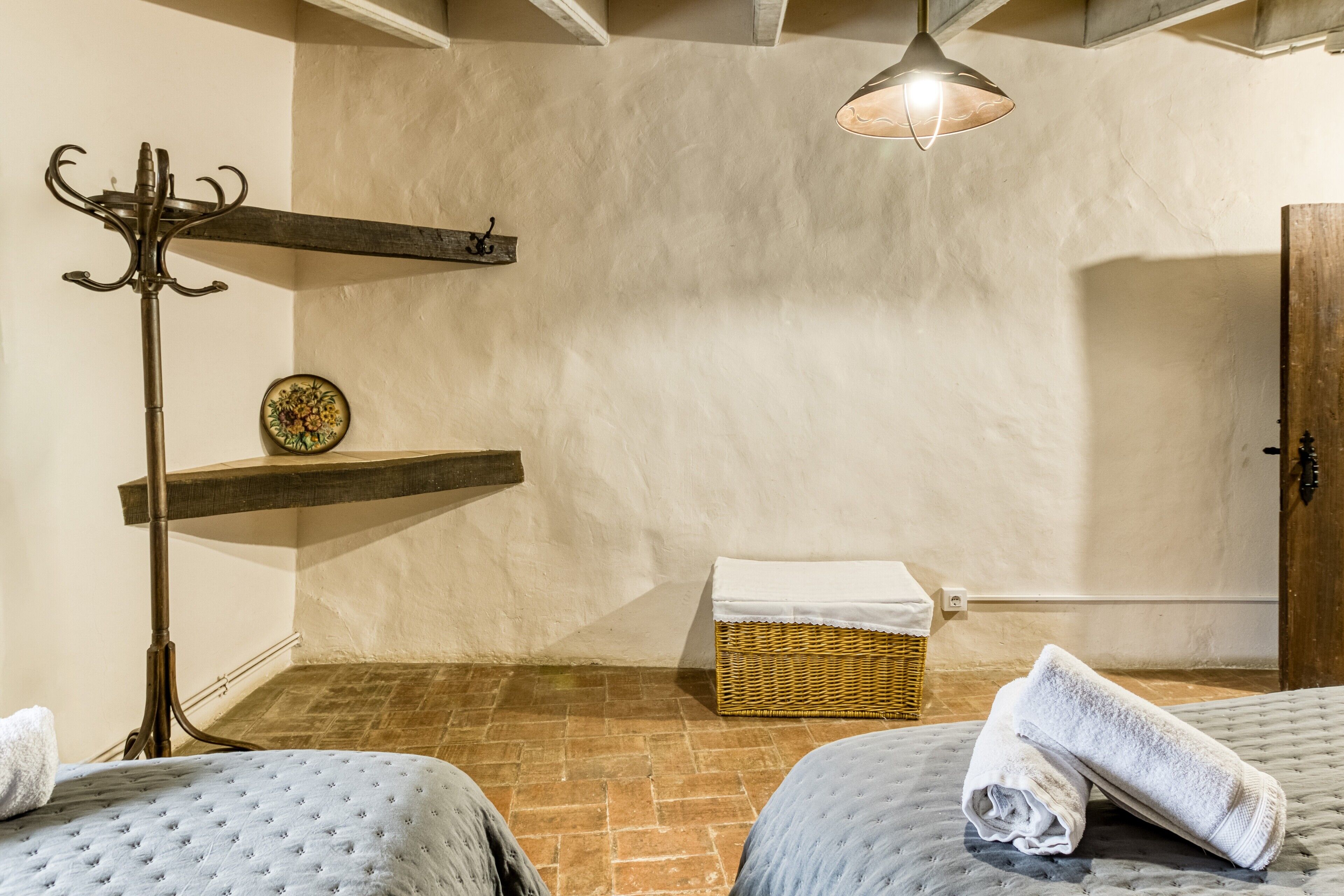 Una habitación con dos camas, un estante de madera, una cesta de mimbre, un perchero y una lámpara colgante.