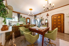 A dining area with a wooden table and green chairs, a kitchen in the background, and a large wooden door.