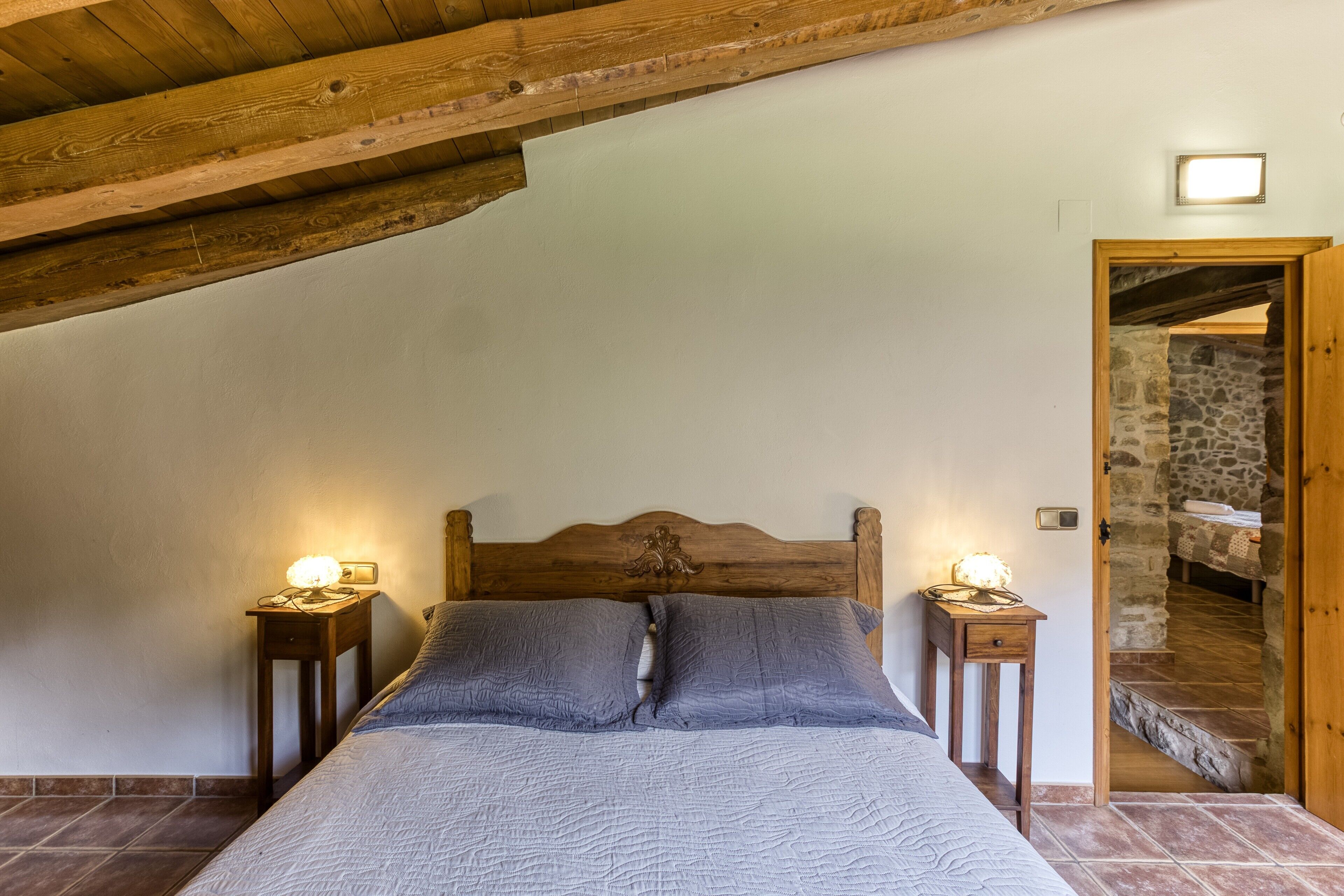 A bedroom with a wooden ceiling, a bed with grey bedding, two bedside tables with lamps, and a stone wall.