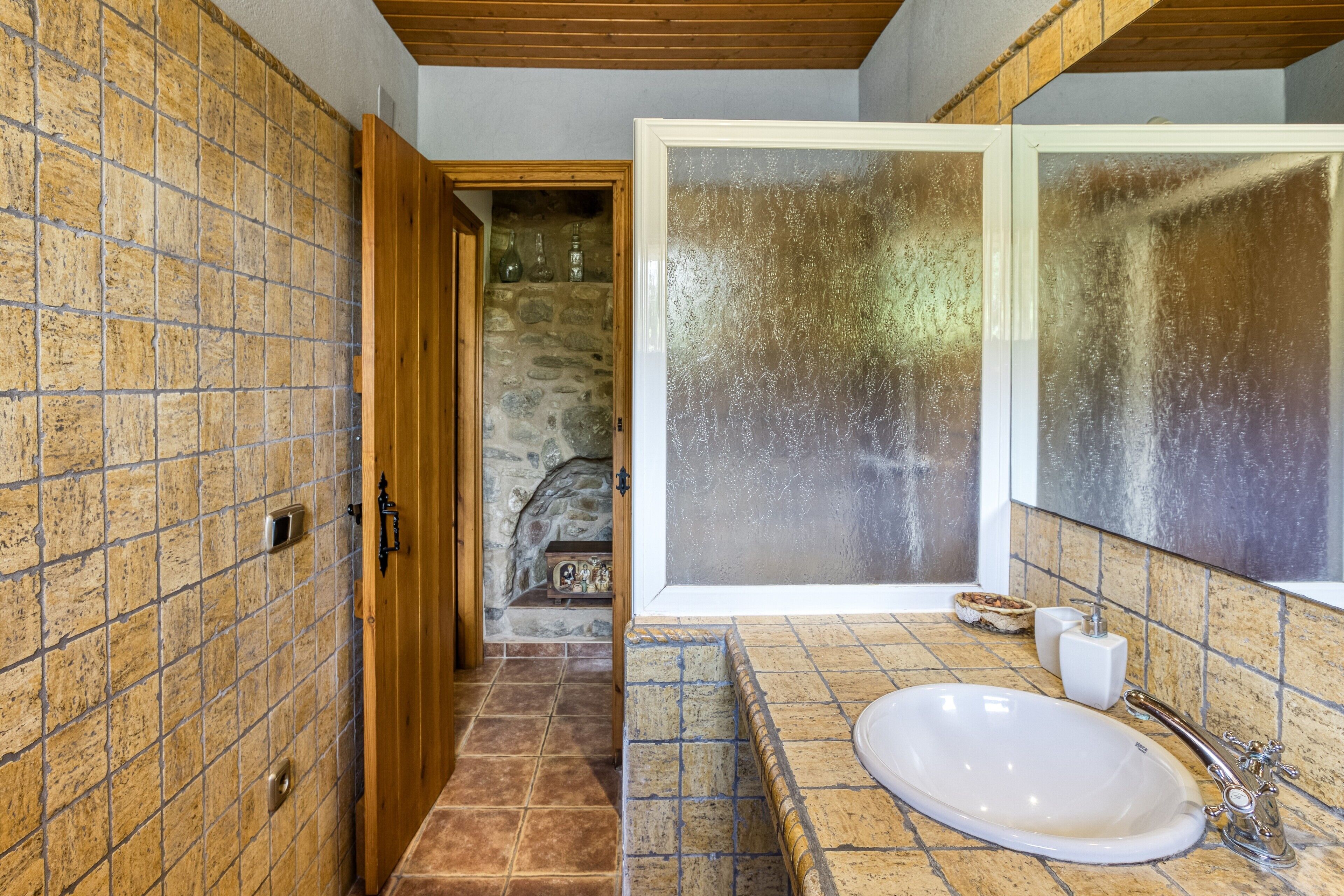 Ein Badezimmer mit Steinwand, Holztür, Fliesenboden, Waschbecken mit Spiegel und Fenster.