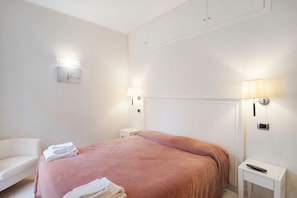 A hotel room with a bed, two bedside tables, a nightstand, a chair, and a wall-mounted light fixture.